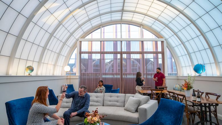 The image shows a bright room with a curved ceiling, people conversing on couches, and standing at a table, featuring globes and modern decor.