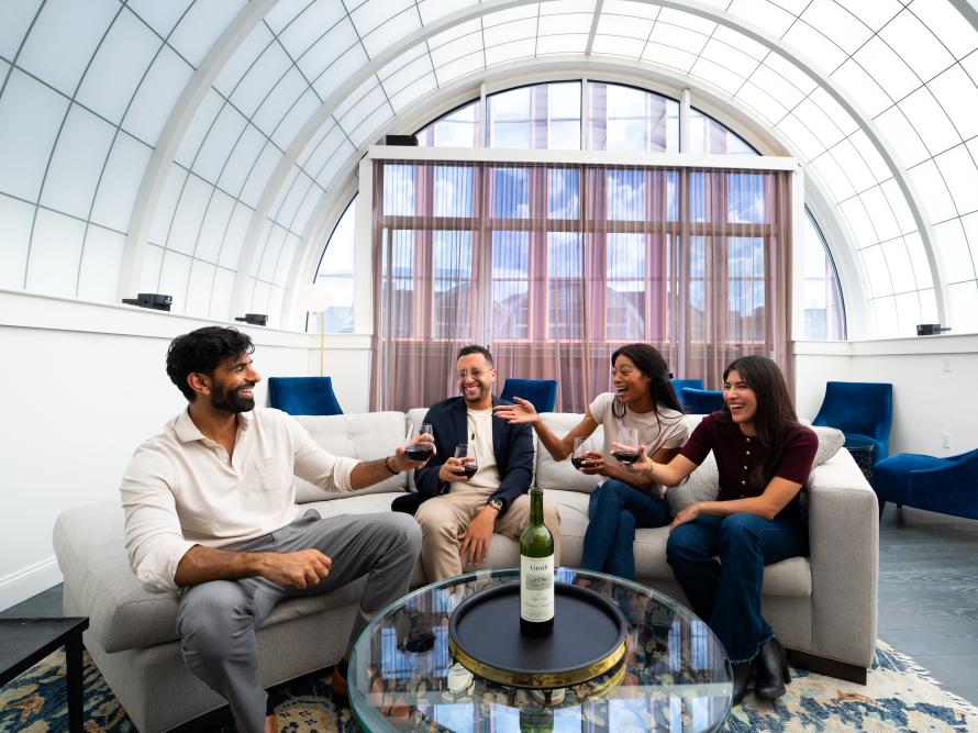 A group of friends cheers with wine in a bright lounge with blue chairs and a glass table, under a vaulted window.