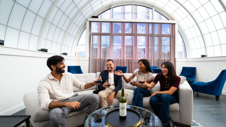 A group of friends cheers with wine in a bright lounge with blue chairs and a glass table, under a vaulted window.