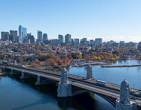 A city skyline with a bridge stretching over a river, surrounded by trees and buildings under a clear blue sky.