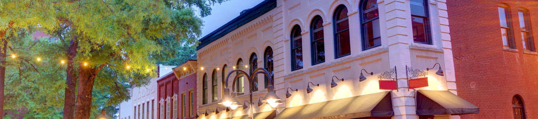 Charming streetscape with historic brick buildings, arched windows, and decorative lights under a large tree.