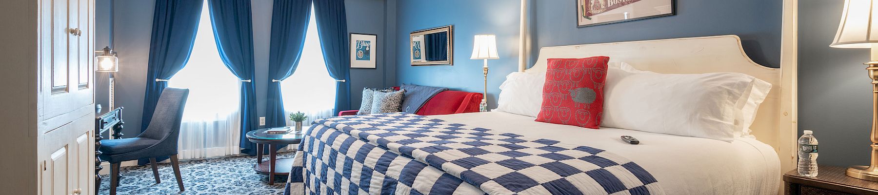 A stylish bedroom features a large bed with a checkered quilt, elegant blue curtains, cozy seating, and warm lighting for a relaxing atmosphere.