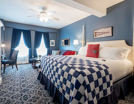 A stylish bedroom features a large bed with a checkered quilt, elegant blue curtains, cozy seating, and warm lighting for a relaxing atmosphere.