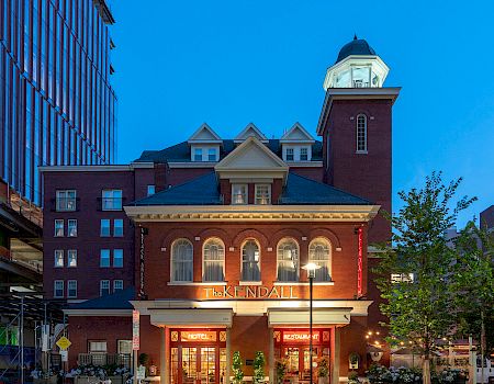 A historic brick building with illuminated windows and a tower, set against a clear blue sky, flanked by modern buildings.