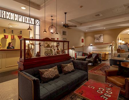The image shows a warmly lit hotel lobby with a reception area, cozy seating, vintage decor, and a relaxed atmosphere.