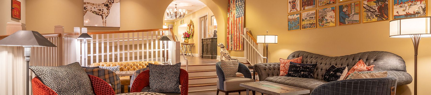 A cozy living room with patterned rugs, armchairs, a sofa, wall art, lamps, and a stairway leading to another room.