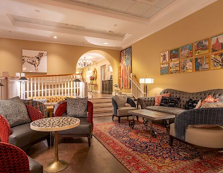 A cozy living room with patterned rugs, armchairs, a sofa, wall art, lamps, and a stairway leading to another room.