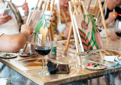 People are painting on canvases at easels with wine glasses and palettes on the table, creating a lively, artistic atmosphere.