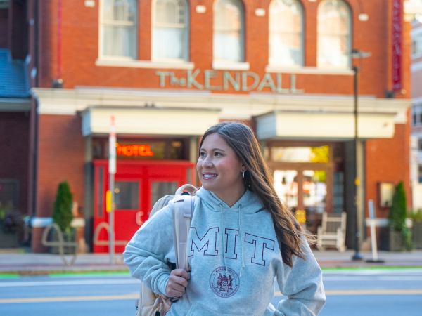 A person in an MIT sweatshirt walking on a street in front of a brick building labeled "TK KENDALL."