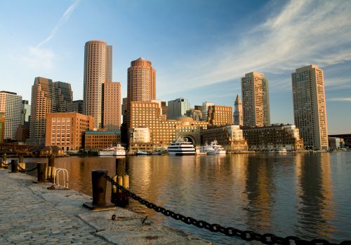 The image shows a waterfront city skyline with tall buildings reflecting in the calm water, and boats docked along the shore in the foreground.