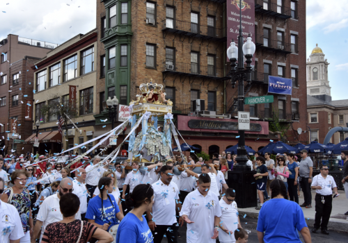 A crowded street scene with people in white shirts and a religious procession carrying a statue decorated with streamers.