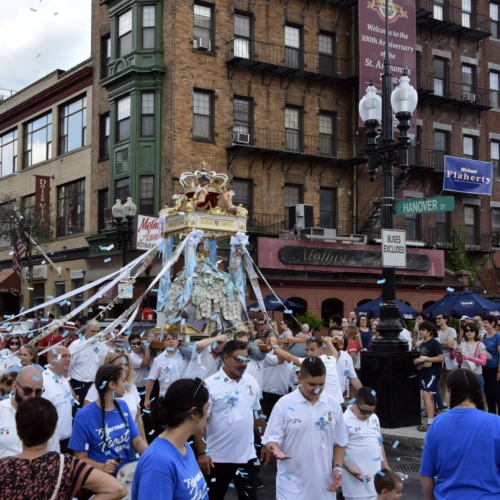 A crowded street scene with people in white shirts and a religious procession carrying a statue decorated with streamers.