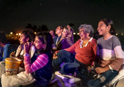 A group of people enjoying a movie outdoors at night, sitting on blankets and sharing popcorn, smiling and watching intently.