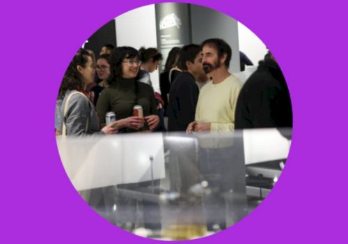 People are gathered at an event or exhibition, with some holding drinks, set against a purple circular frame.