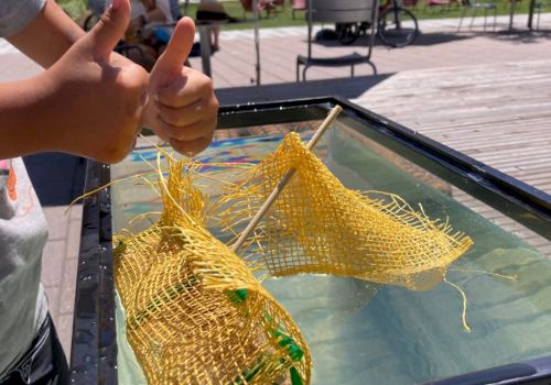 A child gives thumbs up near a water tank with yellow nets stretched on rods, outdoors.