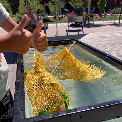 A child gives thumbs up near a water tank with yellow nets stretched on rods, outdoors.