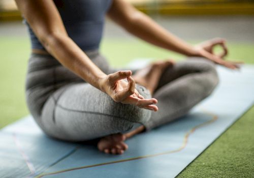 A person is sitting cross-legged on a yoga mat, practicing meditation with fingers in a mudra pose, indoors on a green surface.