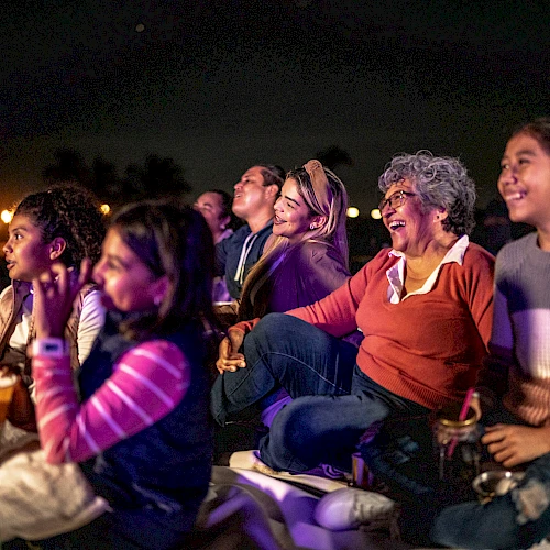 A group of people enjoying a movie outdoors at night, sitting on blankets and sharing popcorn, smiling and watching intently.