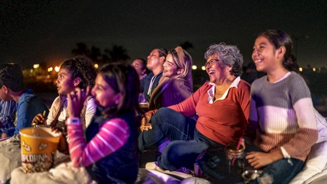 A group of people enjoying a movie outdoors at night, sitting on blankets and sharing popcorn, smiling and watching intently.