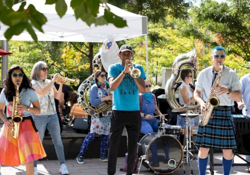 A lively band performs outdoors with colorful costumes and various instruments, creating a festive atmosphere under a white canopy.