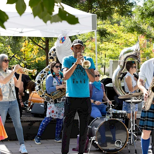 A lively band performs outdoors with colorful costumes and various instruments, creating a festive atmosphere under a white canopy.