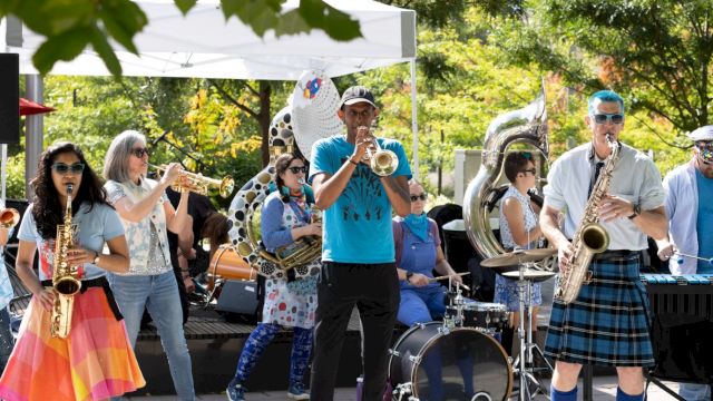 A lively band performs outdoors with colorful costumes and various instruments, creating a festive atmosphere under a white canopy.