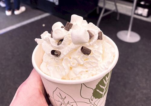 A paper cup filled with whipped cream, marshmallows, and chocolate chips. It is being held by a hand in an indoor setting.