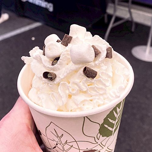 A paper cup filled with whipped cream, marshmallows, and chocolate chips. It is being held by a hand in an indoor setting.