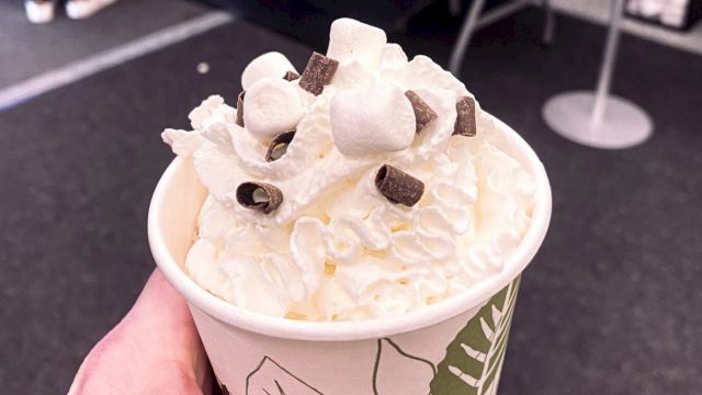 A paper cup filled with whipped cream, marshmallows, and chocolate chips. It is being held by a hand in an indoor setting.