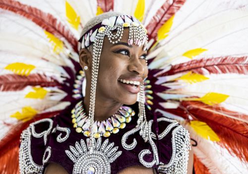 A person in vibrant, ornate costume with feathers and beads, smiling and looking to the side, set against a colorful, festive background.