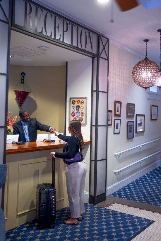A reception area with a person checking in at the desk. The receptionist is assisting, and there's a suitcase on the floor.