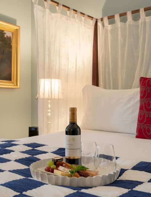 A cozy room features a patterned bedspread, pillows, wine with glasses on a tray, a framed painting, and a vintage mirror on the wall.