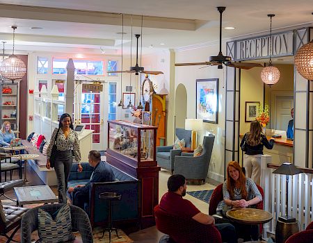 The image shows a cozy interior with people sitting and walking around, near a reception area. The space has warm lighting and comfortable seating.