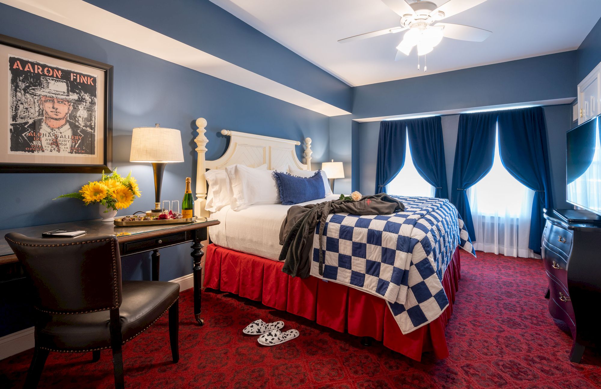 A cozy hotel room with a double bed, checkered blanket, desk with a chair, lamp, sunflowers, and artwork on the blue walls.