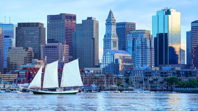 A sailboat is on the water with a city skyline in the background, featuring modern skyscrapers and an iconic clock tower.