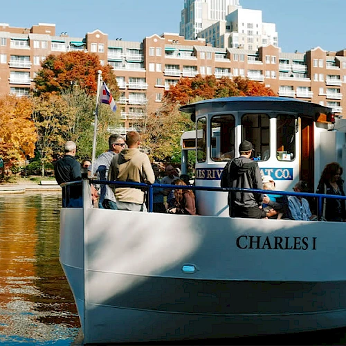 A small boat named "Charles I" with passengers onboard in front of a riverside with trees and buildings in the background.