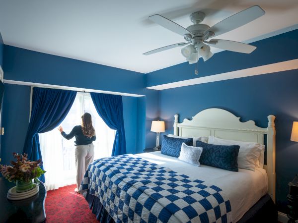 A person stands near a window in a blue-themed bedroom with a checkered bedspread and ceiling fan.