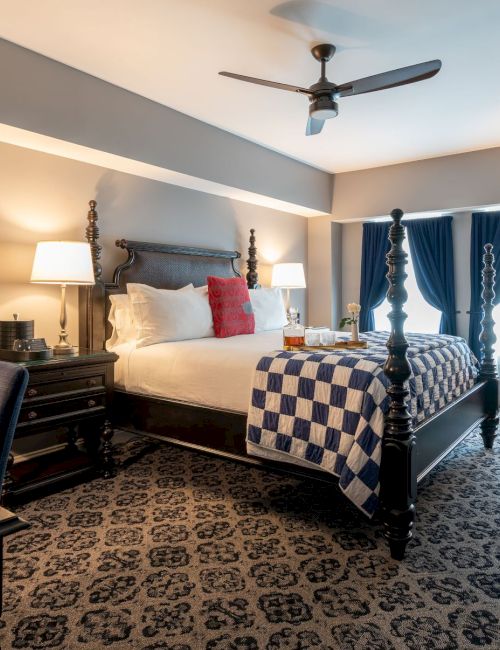 The image shows a cozy bedroom with a large bed, carpeted floor, patterned chair, desk, lamps, and a ceiling fan, creating a warm atmosphere.