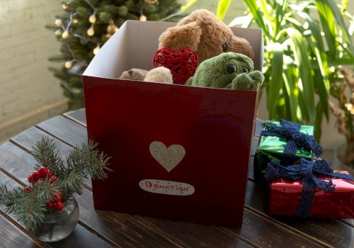 The image shows a red gift box with stuffed toys, a decorated plant, and wrapped presents on a wooden table.