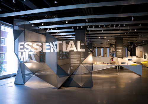 A modern gallery or showroom with angular gray geometric partitions spelling “ESSENTIAL MIT” across a bright, open space with displays and seating in the background.