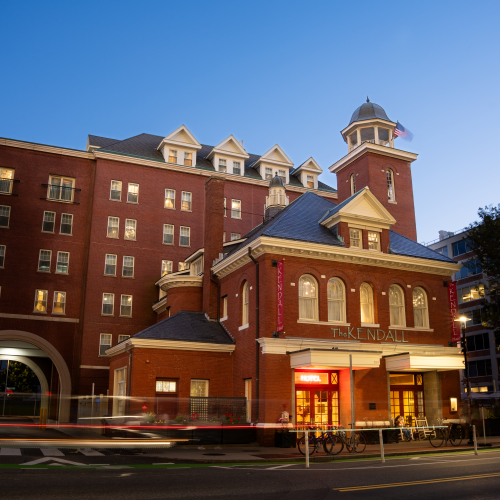 A brick hotel-like building with a tower, arched windows, and bright ground-floor storefronts along a street at dusk.