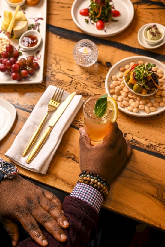 A gourmet table with cheeses, grapes, chickpeas, and small plates; a hand holds a drink as gold utensils and a napkin set a stylish scene.