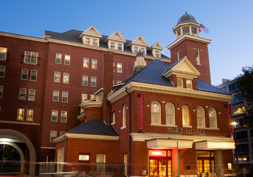 A brick hotel or historic building with a blue roof, tower, and bright storefront at street level, under a clear blue evening sky.
