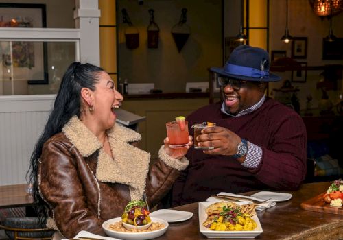 Two friends laughing happily over cocktails and plates of colorful food at an outdoor restaurant.