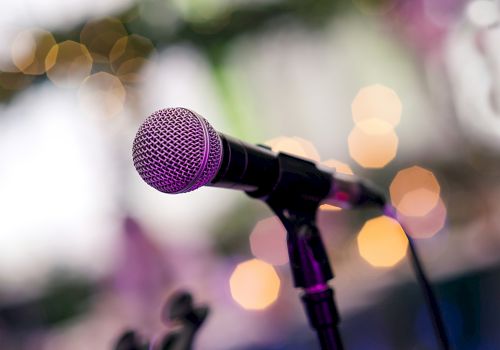 A close-up of a purple microphone on a stand, set against a blurred colorful stage backdrop with warm bokeh lights, likely at a performance.
