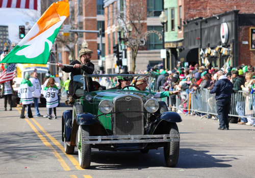 A vintage car parade drives down a city street as a man waves a large Indian flag; spectators line the sidewalks behind barriers.