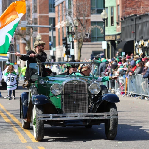 A vintage car parade drives down a city street as a man waves a large Indian flag; spectators line the sidewalks behind barriers.