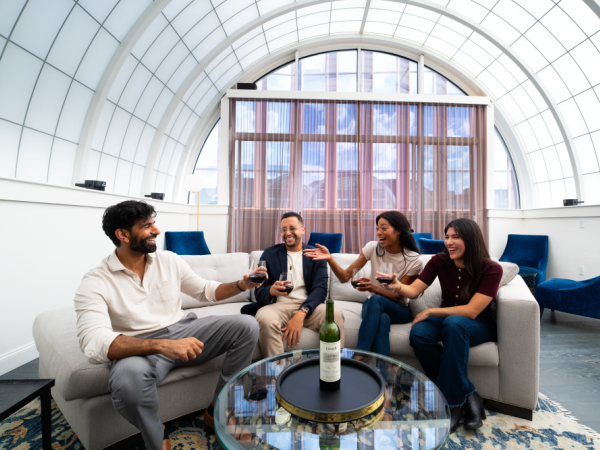 A group of four friends sits in a bright lounge, toasting with wine glasses around a glass coffee table&mdash;sunlit arched window backdrop, modern blue chairs.