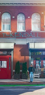 The KendalL storefront with red door, brick building, and a person walking on the sidewalk at street level.