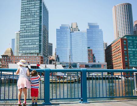 Two children stand at a blue railing by a waterfront, facing skyscrapers and boats in a sunny city harbor.